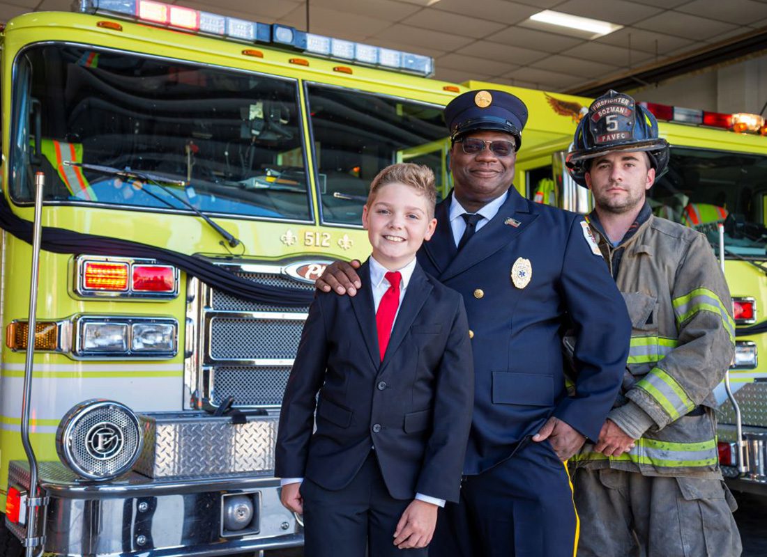About Our Agency - Young Man with Suit on Smiling and Standing with an Officer and Firefighter in Front of a Yellow Firetruck